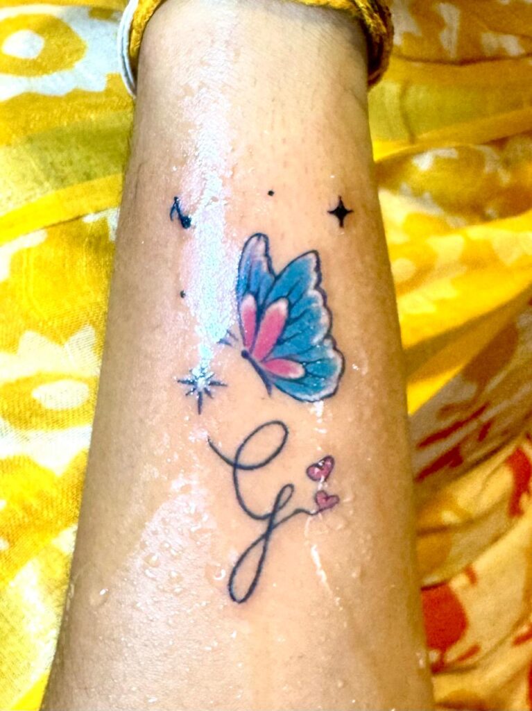 A close-up tattoo featuring a blue pink butterfly, musical note and a sparkle around it, there is a cursive letter “G” ending with two small pink hearts, all inked on the forearm.