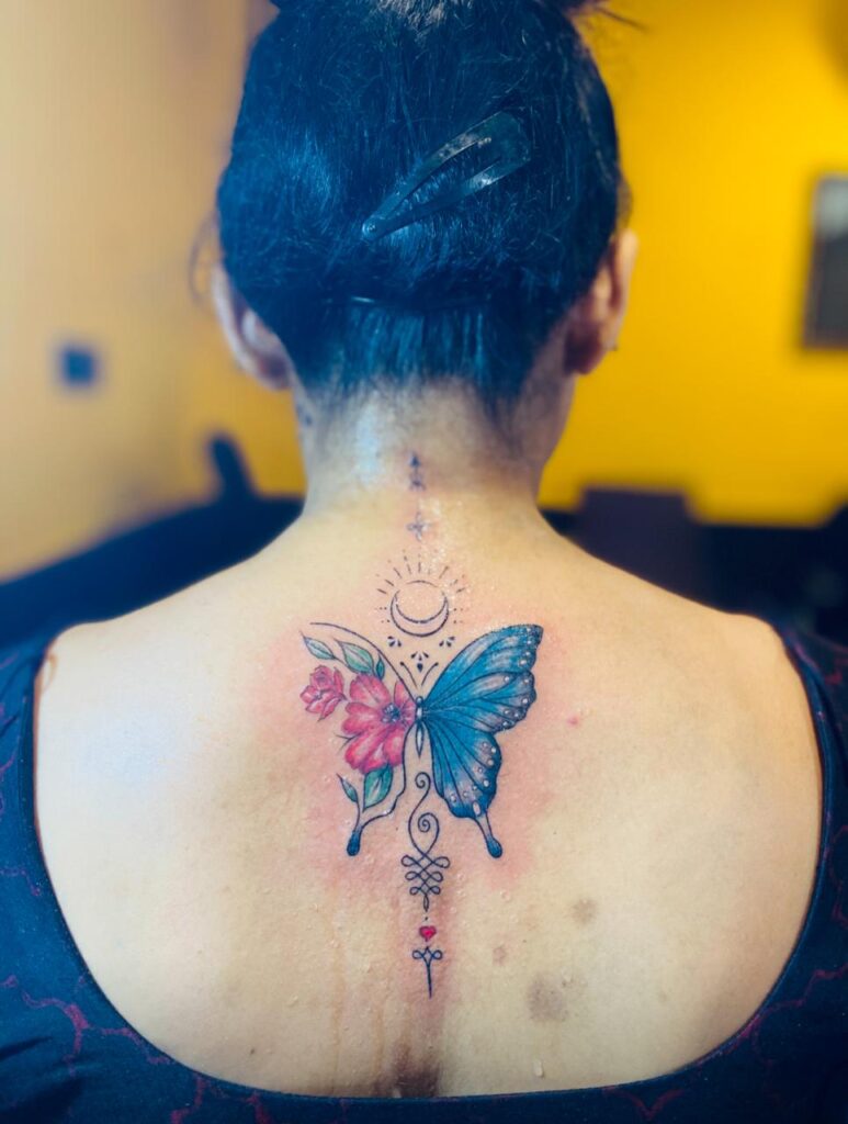 The image shows a colorful upper back tattoo featuring a half-butterfly, half- floral design with a crescent sun-moon symbol at the top of it and unalome ending in a red heart below.