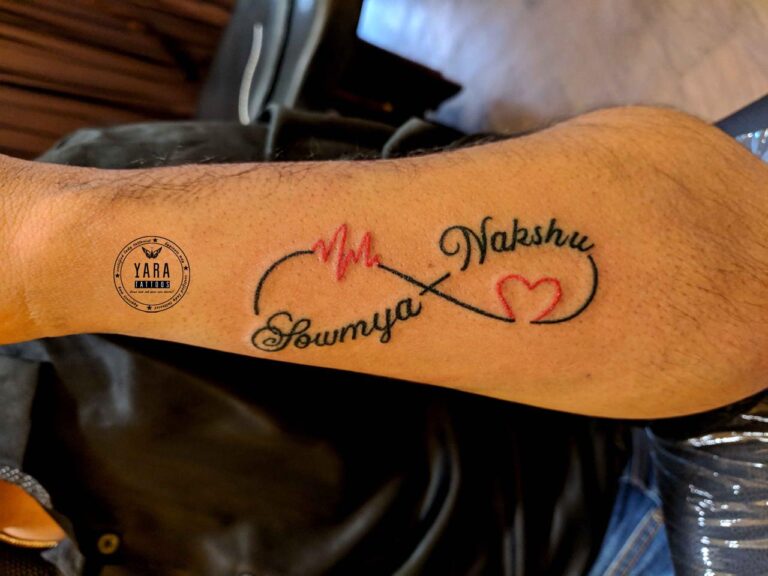 The image shows the tattoo “Sowmya” is written on the left of the infinite loop and the name “Nakshu” is written on the right of the loop along with a red heart and heart beat symbol.