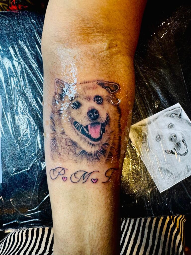 The image shows a realistic dog portrait tattoo on a person’s arm. Below that portrait the initials P M I were written in elegant cursive form, each separated by a pink heart.
