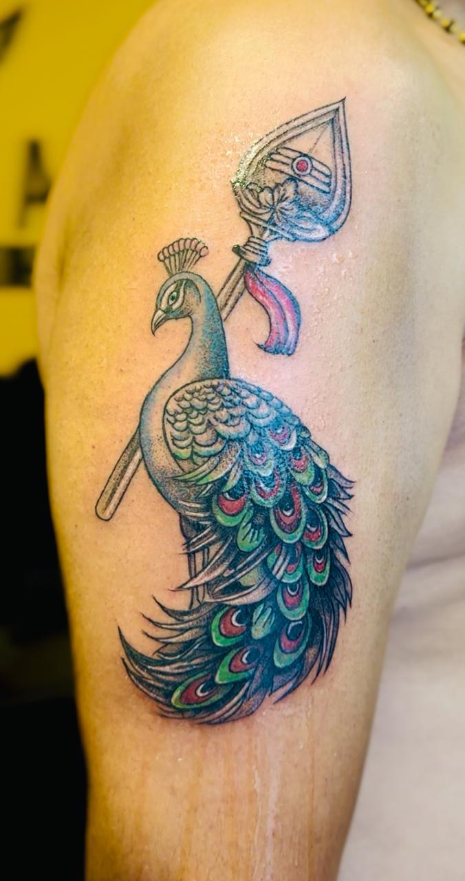 The image displays a tattoo featuring a peacock and Murugan’s Vel.