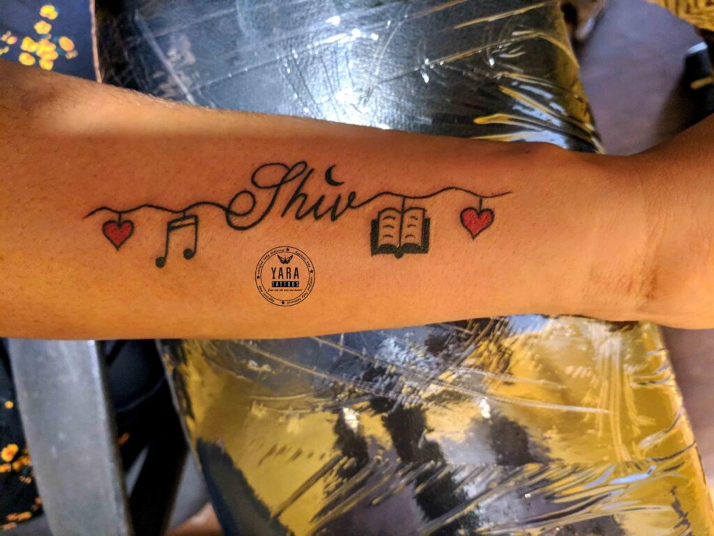 The image shows a tattoo on the forearm featuring the name “Shiv” with a crescent on the top of “i” instead of a dot at the centre connects with a thin curvy line like string connecting a small red heart followed by a musical note to the left and an open book with pages and then another small red heart to the right.