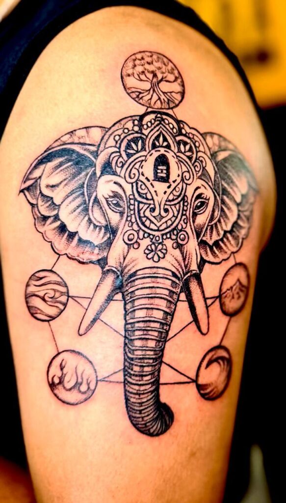 The image shows an intricate blackwork tattoo of an elephant head adorned with mandala-like patterns. Above the elephant there’s a circle containing a tree of life and around it several connected circles representing cosmic elements.