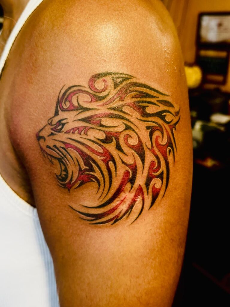 The image shows a vibrant tribal-style lion tattoo with black and red accents on a person’s upper arm. The lion has stylized flowing, flame-like and swirling patterns.