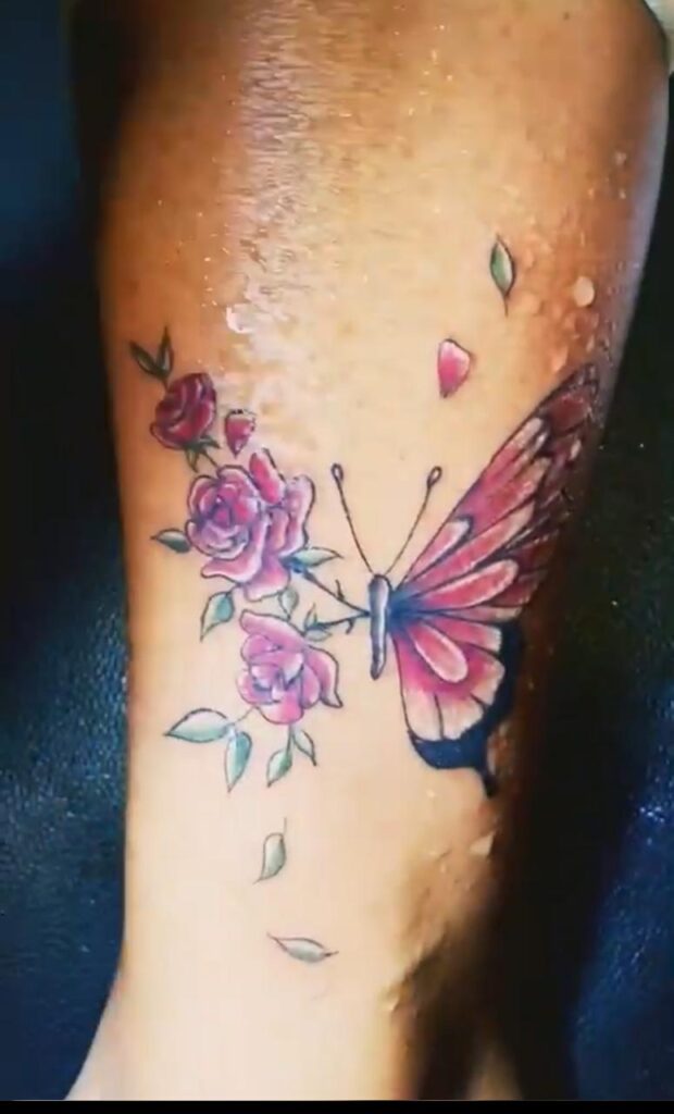 The image shows a butterfly tattoo in red, pink and black shade, on the right with a colourful wing and on the left it contains roses and leaves.