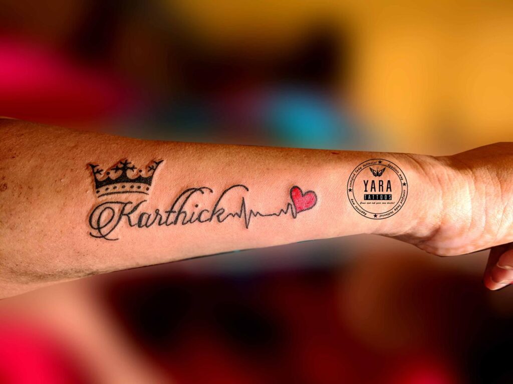 The image features a name tattoo that reads Karthik along with a crown above “K” and the name followed by a heart beat line ends with a red heart.