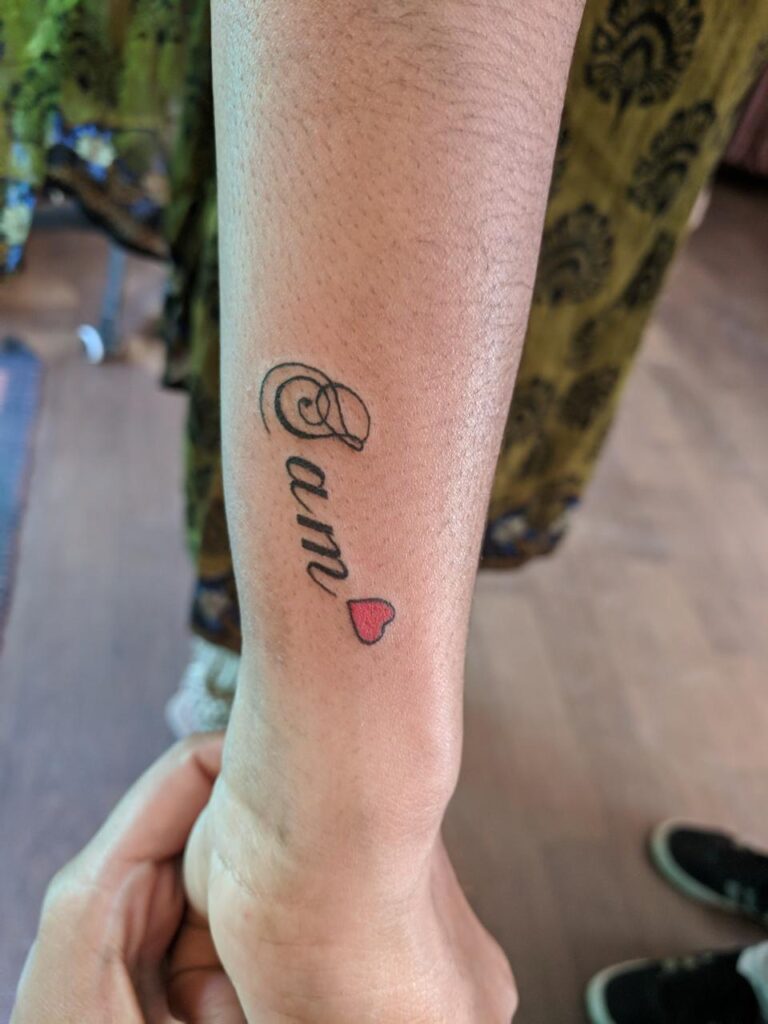 The image shows a name tattoo, “sam” in a simple hand-written style along with a tiny red heart.