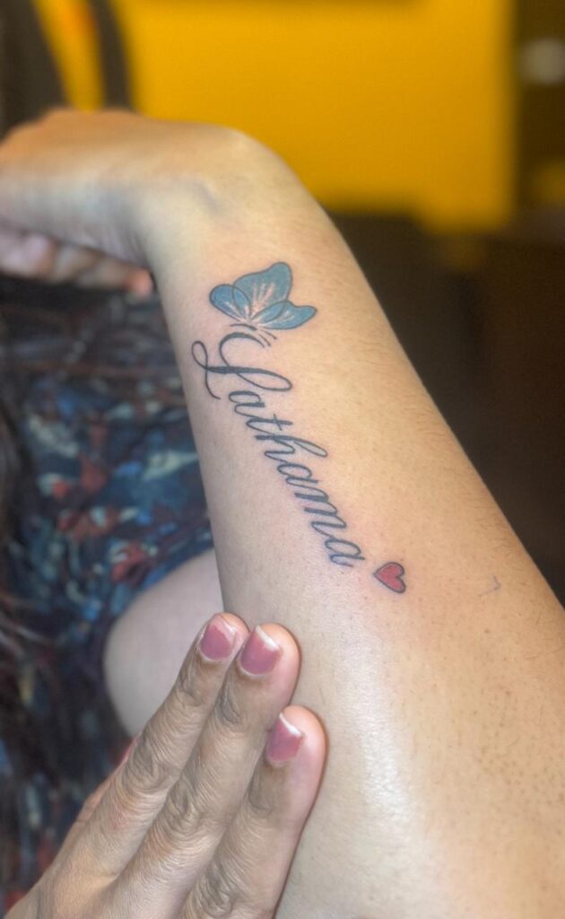 The image shows a tattoo featuring the name “Lathama” along with a cute little blue butterfly on top of letter “L” and a little red heart above “a” at the end.