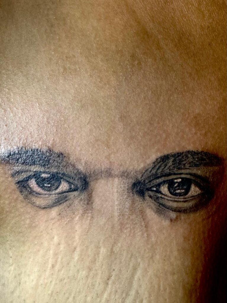 The image shows a black and grey portrait tattoo featuring the eyes of a person in a realistic way.