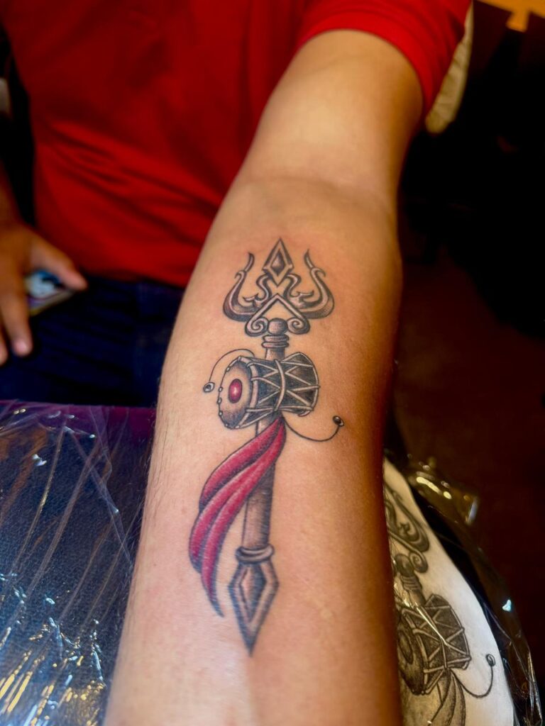The image shows a permanent tattoo showing a stylized trishul design with a damaru in the middle, with a red ribbon wrapped around a pointed spear like end.