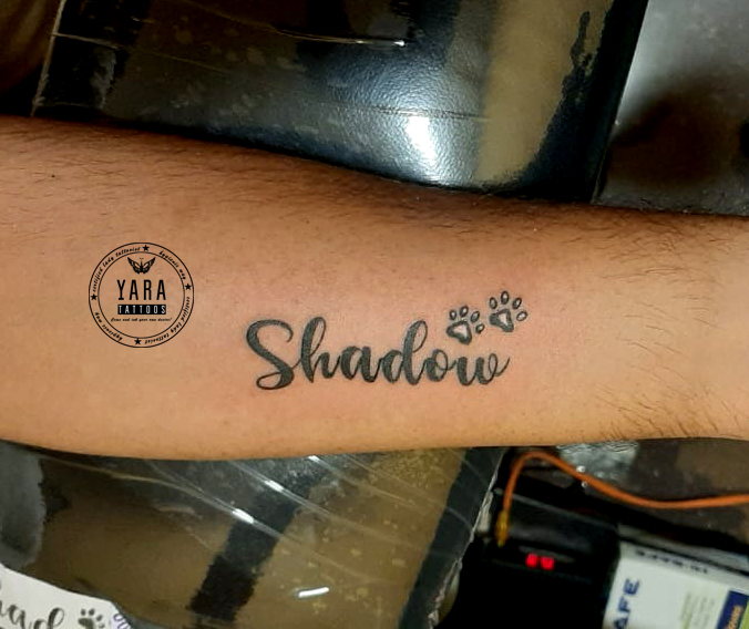 Minimal name tattoo reading “Shadow” in cursive script with small paw prints, inked on the inner forearm in black ink