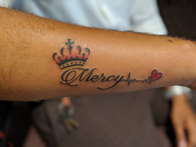 Decorative name tattoo reading “Mercy” in elegant cursive, topped with a small crown and finished with a heartbeat line and red heart, inked on the forearm.