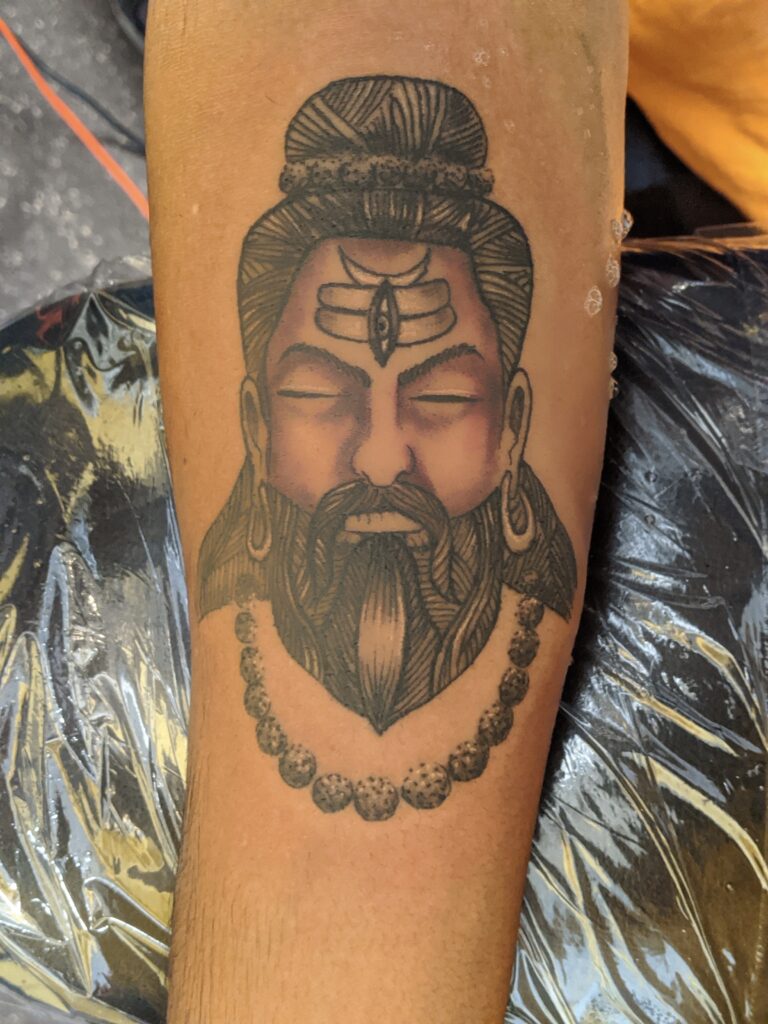 Detailed black and gray portrait tattoo of a meditating sage with closed eyes, topknot hair, sacred forehead markings, and a beaded necklace, inked on the forearm with soft shading.