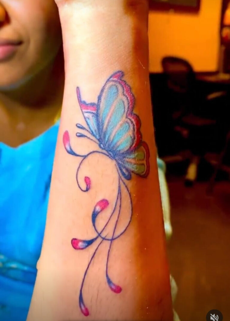 Close-up of a person’s forearm featuring a colorful butterfly tattoo with blue, teal, and pink wings, flowing into elegant, swirling lines that trail down the arm.