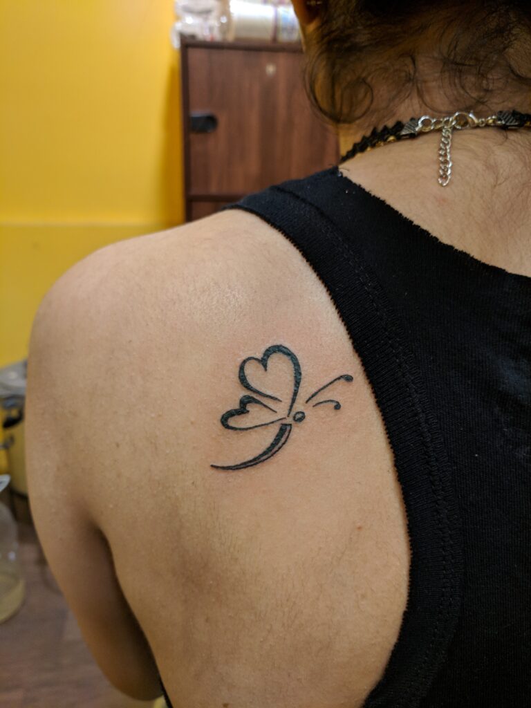 Minimal black butterfly tattoo with heart-shaped wings inked on the upper shoulder